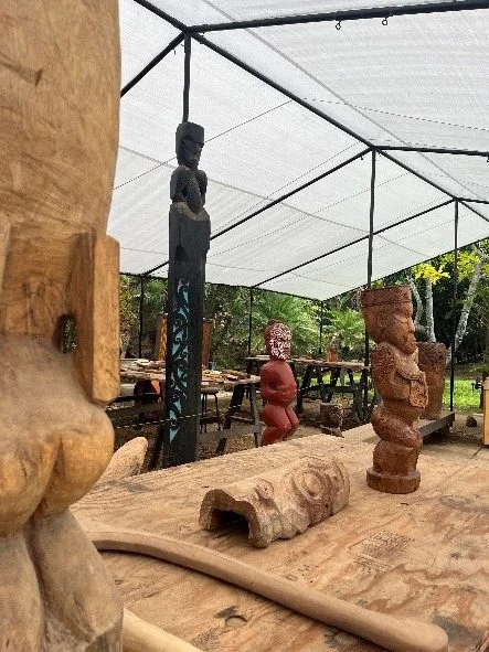 A tented area with work tables. Several hand-carved items are on and around the table in the foreground, including tikis.