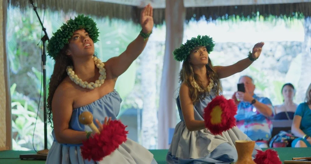 Two female dancers in green wreath crowns and blue Hawaiian costumes demonstrate a hula dance done while kneeling.