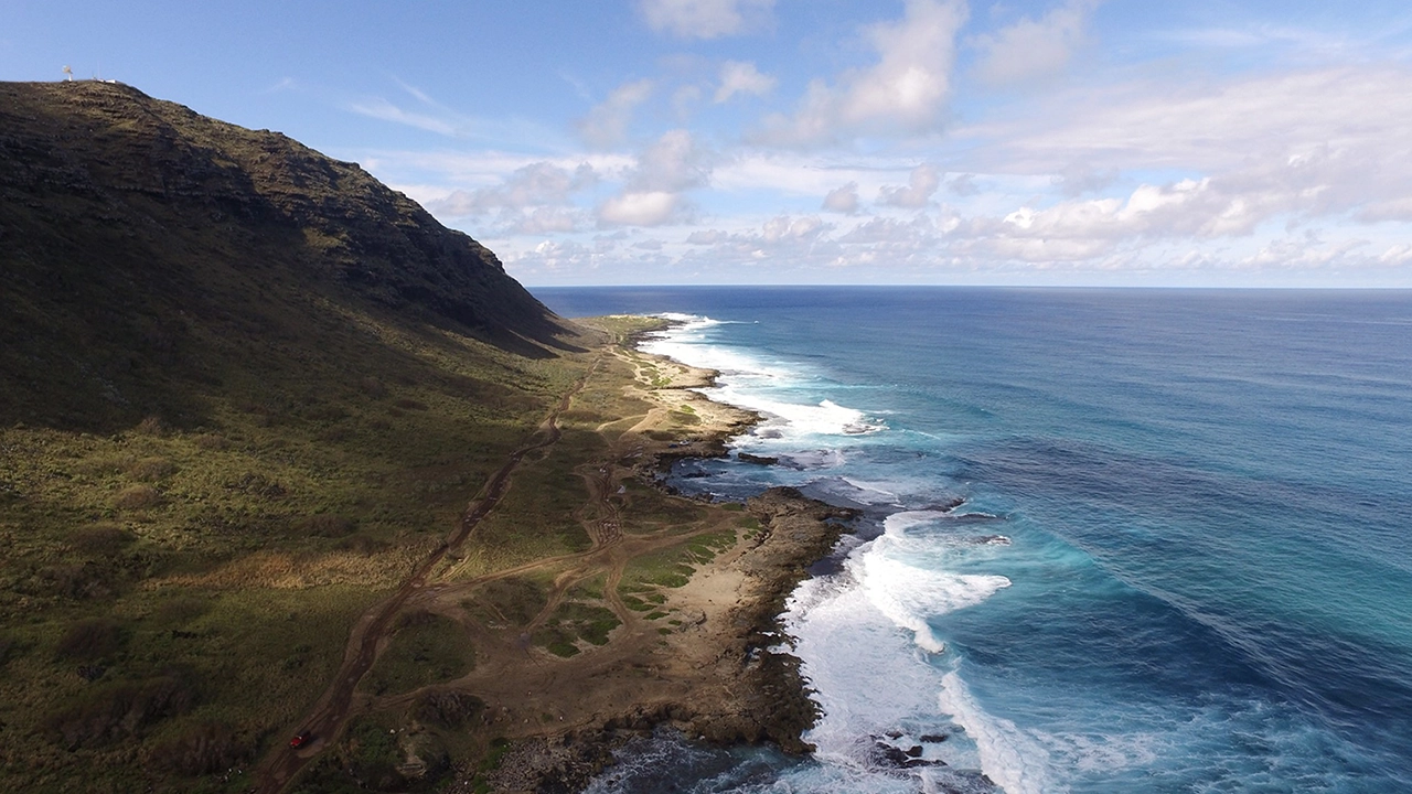 View of rocky island land and deep blue ocean with a towering mountain and low lying shrubbery.