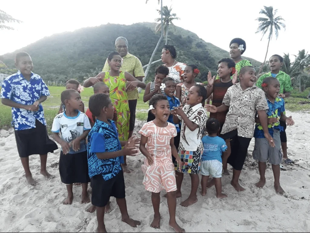 A group of many Fijian men, women, and children, all happily singing on the beach.