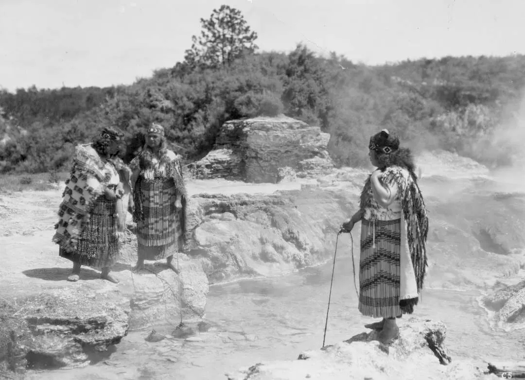 Historical photo of Māori women in traditional clothing, cooking food in flax baskets which have been lowered into hot springs.