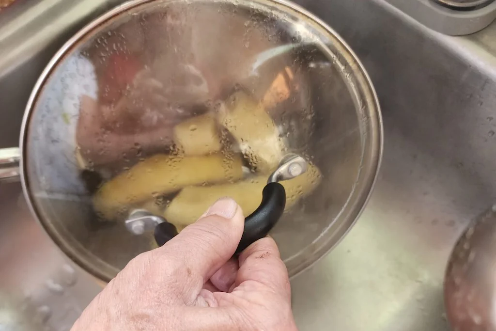 Demonstrating how to drain the boiled bananas by holding the lid on a pot while pouring into a sink.