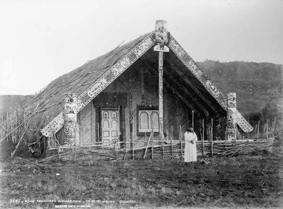 An old photograph of King Tāwhiao's whare, a large, A-frame style building with a thatched roof and intricate Māori designs. 