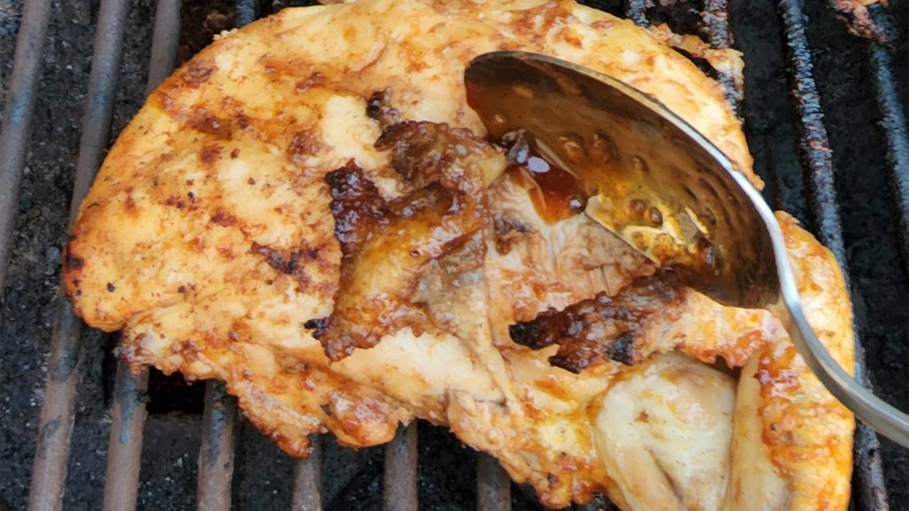 Close-up of a hand using a spoon to brush rich, mahogany-colored Huli Huli glaze onto grilling chicken pieces