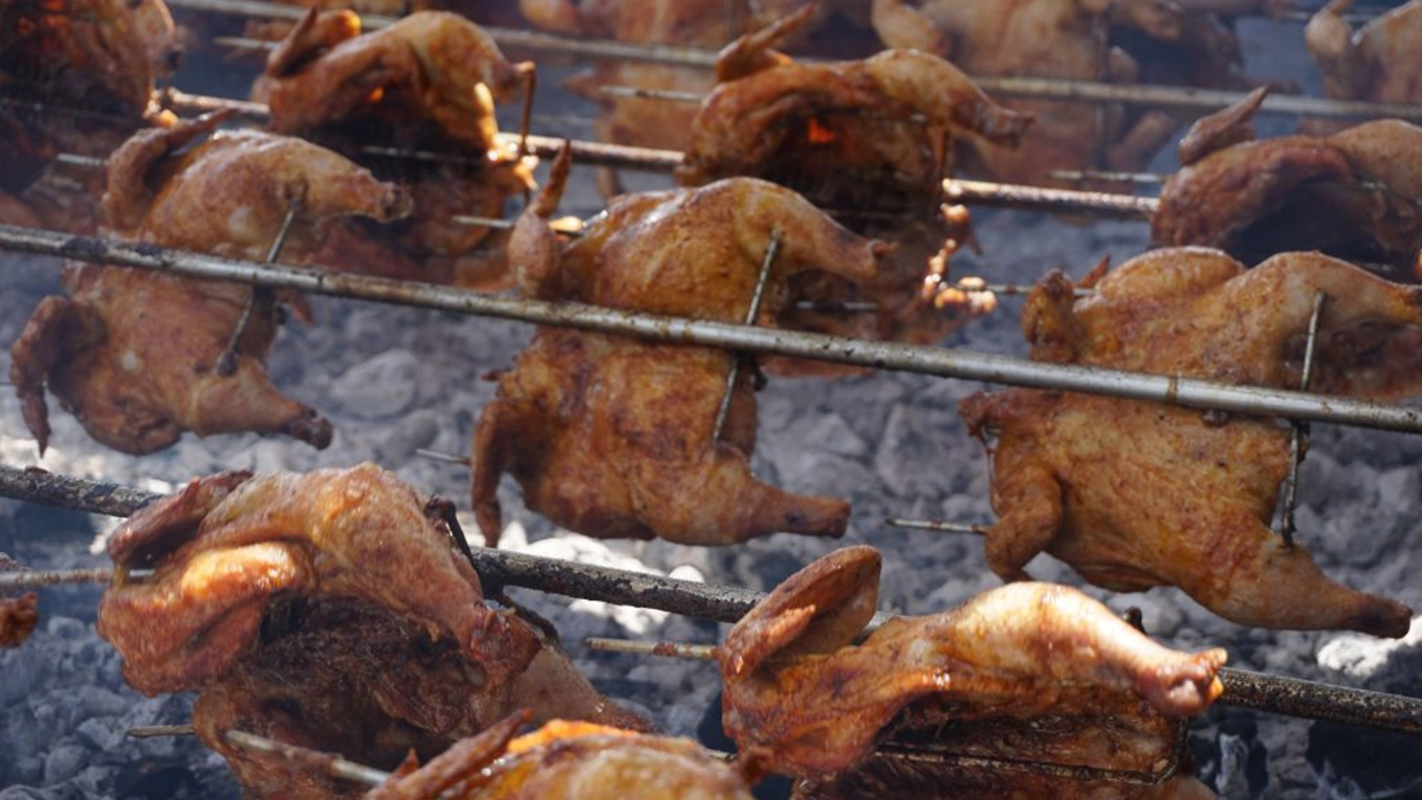 Chicken pieces turning on a rotisserie spit over an open flame, the exterior deeply caramelized from the frequent turning and glazing.