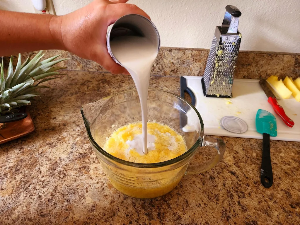 A can of coconut milk being poured into a measuring cup with the grated pineapple.